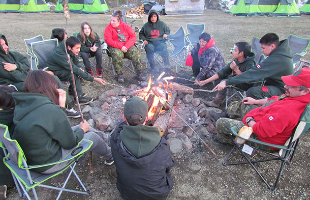 Junior Canadian Rangers chat and cook hot dogs around a camp fire