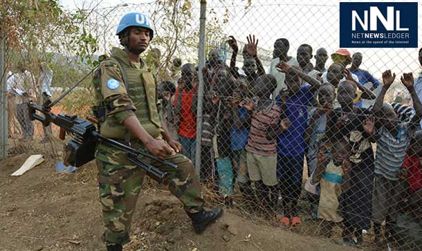 A peacekeeper stands guard at UN House while displaced children look on behind a fence. UN Photo/Isaac Billy