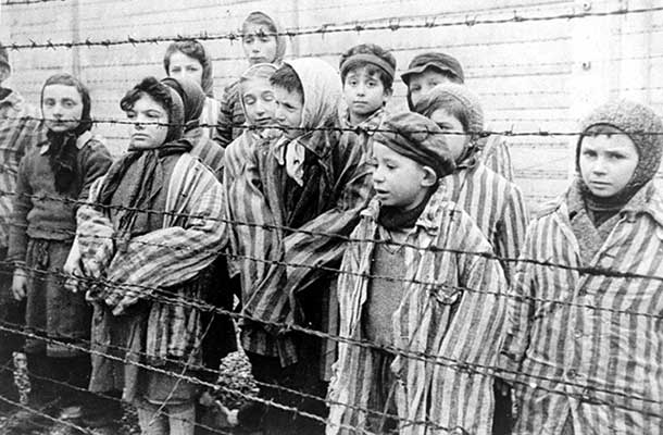 Still photograph from the Soviet Film of the liberation of Auschwitz, taken by the film unit of the First Ukrainian Front, shot over a period of several months beginning on January 27, 1945 by Alexander Voronzow and others in his group. Child survivors of Auschwitz, wearing adult-size prisoner jackets, stand behind a barbed wire fence. Among those pictured are Tomasz Szwarz; Alicja Gruenbaum; Solomon Rozalin; Gita Sztrauss; Wiera Sadler; Marta Wiess; Boro Eksztein; Josef Rozenwaser; Rafael Szlezinger; Gabriel Nejman; Gugiel Appelbaum; Mark Berkowitz (a twin); Pesa Balter; Rut Muszkies (later Webber); Miriam Friedman; and twins Miriam Mozes and Eva Mozes wearing knitted hats.