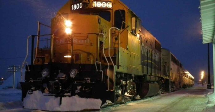 Ontario Northlander parked at a train station during twilight in a snowy setting. The locomotive's headlights and station lighting cast a warm glow on the surrounding area, contrasting with the blue hues of the early evening sky