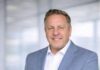 Jody Humphrey Redefines the Role of Financial Education in Today’s Economy Professional man in a light blue blazer and white shirt posing confidently in a bright office hallway, hands in pockets.